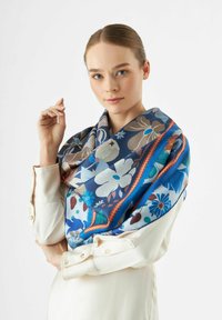Écharpe en soie avec des motifs floraux dans des tons de bleu, présentant un motif vibrant avec des accents de rouge et de marron. Portée drapée sur les épaules.