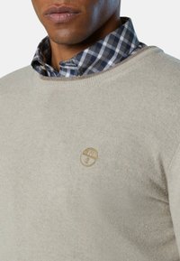 Maglione crema con scollatura rotonda e polsini a costine. Indossato su una camicia a quadri blu e bianca. Presenta un piccolo logo ricamato.