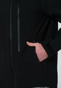 Svart zip-up jacka tillverkad av slätt tyg. Har två bröstfickor och en sidficka med eleganta dragkedjor och kardborreaccent vid ärmsluten.