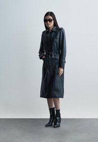 Zwarte leren trenchcoat met knoopsluiting aan de voorkant en ceintuur in de taille, voorzien van een kraag en twee zakken. Het model draagt zwarte enkellaarzen en een zonnebril.