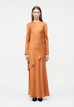 Frau steht, trägt ein langärmeliges, bodenlanges Kleid in verbranntem Orange mit asymmetrischem Lagen-Design, schwarze spitze Schuhe und minimale Ohrringe.