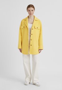 Gelber Wollmischung Oversize-Mantel mit zwei Vorderaschen, goldenen Knöpfen und einem Reverskragen, getragen über einem weißen Top und beigen Hosen.