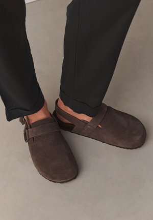 Person trägt dunkelbraune Wildleder-Slipper-Clogs mit Fersenriemen, kombiniert mit schwarzen, aufgekrempelten Hosen auf einem hellgrauen Boden.