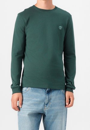 Mann trägt ein dunkelgrünes Langarmhemd mit einem kleinen Baum-Logo auf der Brust und hellblaue Jeans, steht vor einem schlichten Hintergrund.