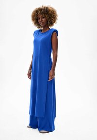 Długa, bezrękawna sukienka w jasnoniebieskim materiale o luźnym kroju, z bocznymi rozcięciami i okrągłym dekoltem; w komplecie z szerokimi, dopasowanymi spodniami.