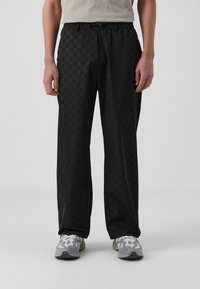 Schwarze gemusterte Hose mit einem elastischen Bund und seitlichen Taschen, aus einem glatten, leichten Stoff gefertigt. Kombiniert mit grauen Sportschuhen.