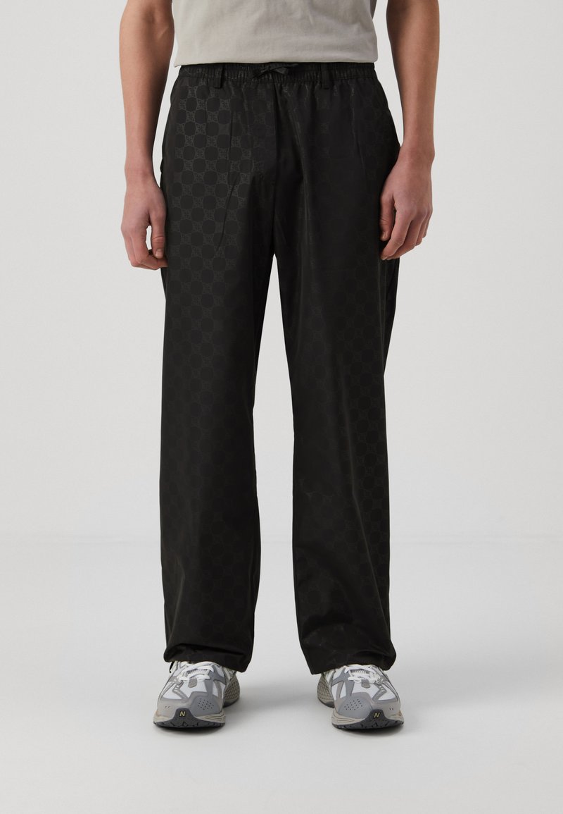 Schwarze gemusterte Hose mit einem elastischen Bund und seitlichen Taschen, aus einem glatten, leichten Stoff gefertigt. Kombiniert mit grauen Sportschuhen.