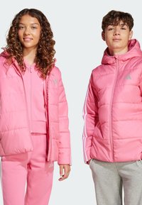 Zwei junge Modelle tragen passende rosa Jacken. Das Mädchen links hat lockiges Haar und trägt ein rosa Outfit, während der Junge rechts kurzes Haar hat und graue Hosen trägt.