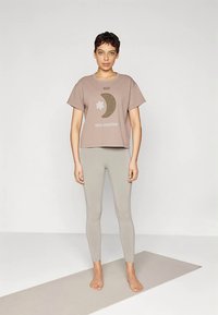 Pruun lühikeste varrukatega T-särk, millel on kuu ja lille graafika; kombineeritud heledate hallide retuusidega. Modell seisab paljajalu jooga mati peal.