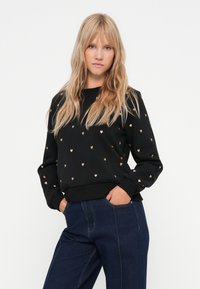 Musthave tumedam, millel on väikesed kuldsed südame mustrid, ümmargune kaelus, soonikud mansettides ning pehme tekstuur, koos tumesiniste teksadega.