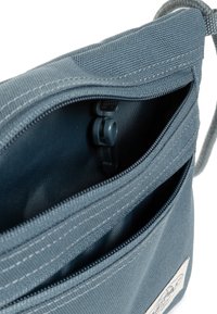 Borsa di stoffa blu con due tasche con zip. Presenta una superficie texturizzata, cuciture a contrasto e una toppa rotonda con logo sul davanti.