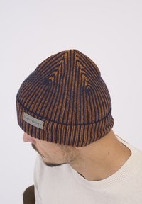 Knitted beanie in navy and orange stripes, featuring a labeled gray patch and a tapered design. Soft texture, suitable for cold weather.
