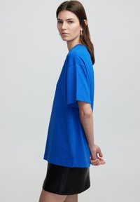 Blauwe katoenen t-shirt met korte mouwen, losse pasvorm, ronde halslijn en zichtbare stikdetails. Gdraagt met een zwarte leren rok.