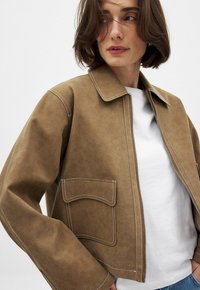 Giacca in suede marrone con colletto a camicia, zip frontale e dettagli in cucitura bianca. Presenta una tasca sul petto e una vestibilità comoda.