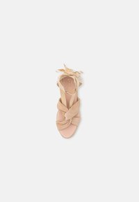 Sandal com design em camurça bege e nó, tira ajustável no tornozelo e parte superior texturizada. Sola plana com detalhes trançados perto do calcanhar.