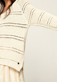 Cream-colored knitted sweater with open weave, horizontal stripes, and side slits. Features a ribbed hem and a small logo detail on the side.