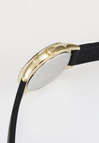 Montre analogique avec un boîtier en acier inoxydable doré, un bracelet en silicone noir, de forme ronde et avec une surface texturée sur le bracelet.