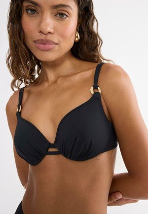 Woman wearing black bikini top with gold ring details on straps and geometric cut-out at center, with wavy brown hair and gold earrings.