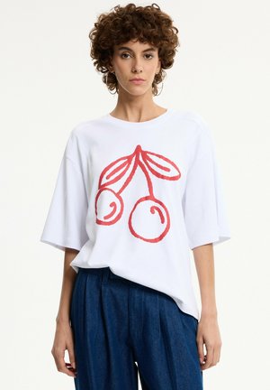 Mujer con cabello corto y rizado, vestida con una camisa blanca oversize con un gráfico de cerezas rojas y pantalones azules de cintura alta sobre fondo blanco.