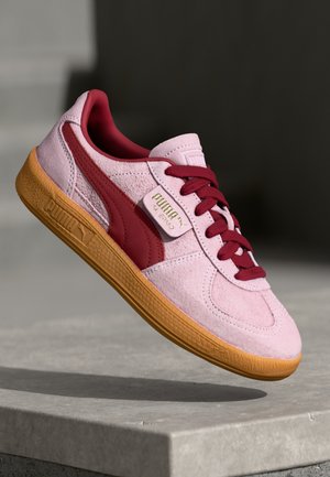 Lyserød ruskindssneaker med røde accenter og brun gummisål, vist midt i luften over en betonoverflade.