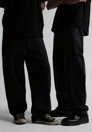 To personer iført løstsiddende sorte bukser og sorte toppe, den ene med sorte sneakers, den anden med sorte lædersko, står side om side.