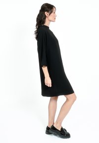 Črna obleka z visokim ovratnikom in ohlapnim krojem, ki ima kratke rokave. Dopolnjujejo jo sijoče črne čevlje z debelim podplatom.