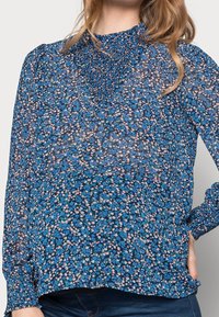 Kvinna som bär en långärmad, blå blommig blus med smockdetaljer vid kragen och ärmsluten, i kombination med mörkblå jeans.