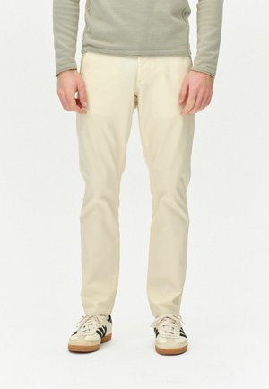 Celana panjang lurus berwarna beige dengan tekstur halus, dipadukan dengan sepatu sneaker putih yang memiliki garis hitam. Atasan yang dikenakan adalah lengan panjang berwarna abu-abu muda.