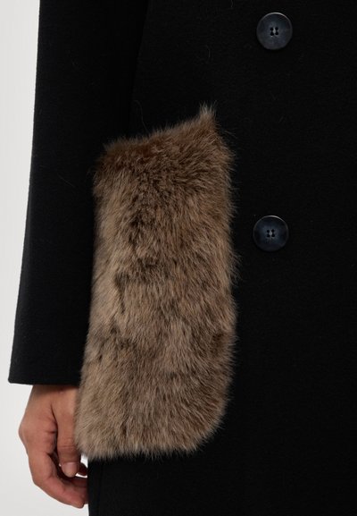 Cappotto nero con una morbida tasca in pelliccia marrone. Il cappotto presenta bottoni neri rotondi e una texture liscia. Una mano tiene la tasca.