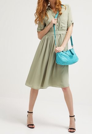 Femme portant une robe midi boutonnée vert olive clair tenant un sac à main turquoise, debout avec les jambes écartées en talons noirs à bride à la cheville.