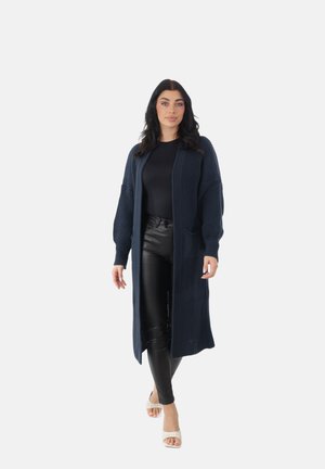 Frau trägt eine lange dunkelblaue Strickjacke, schwarzes Top, schwarze Lederhose und weiße offene High Heels, steht vor einem einfarbigen Hintergrund.