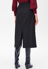 Zwarte midi-jurk met een rechte vorm, een split aan de achterkant en een rits in de taille. Gedragen met hoge, glanzende zwarte hakken.