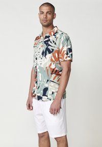 Camisa de manga corta con un patrón de hojas tropicales multicolores, que incluye verdes, cremas y óxido, combinada con pantalones cortos blancos. Textura de tela suave.