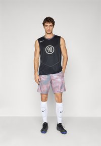 Ung mannlig idrettsutøver iført ermeløs svart drakt med nummer 90, mønstrede shorts, hvite sokker med Nike leggskinn og svarte fotballsko som står.