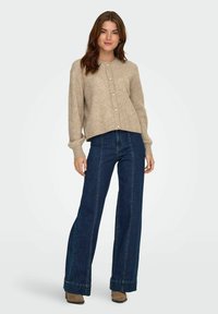 Beżowy sweter dzianinowy z okrągłym dekoltem i perłowymi guzikami, w połączeniu z ciemnoniebieskimi spodniami typu wide-leg. Teksturowana tkanina, swobodny krój.