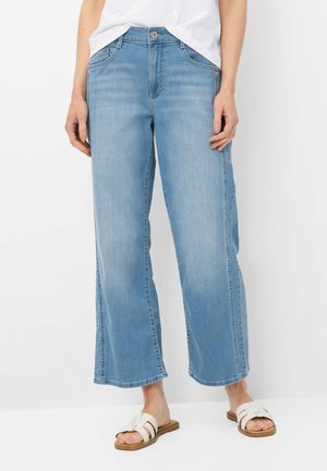 Lichtblauwe rechtvallende jeans, gedragen met een wit topje en witte platte sandalen op een witte achtergrond.