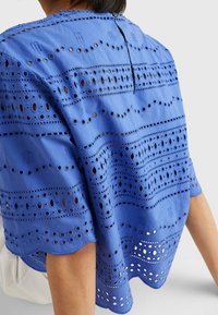 Top blu corto con maniche corte, caratterizzato da motivi ricamati e dettagli a occhio lungo l'orlo e il corpo, con scollatura arrotondata e chiusura a bottoni.