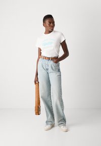 Hvit t-skjorte med korte ermer og blå tekst, lyseblå høyde jeans, brun belte, og hvite sneakers, holder en beige skulderveske.