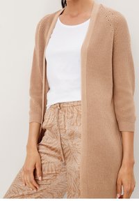 Ljusbeige stickad cardigans med ribbad textur, öppen fram, och tre fjärdedelars ärmar, tillsammans med mönstrade byxor.