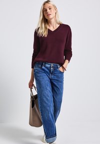 Pull en v Bourgogne, coupe ample, associé à un jean large bleu délavé. Sac fourre-tout beige avec deux poignées. Baskets blanches visibles.