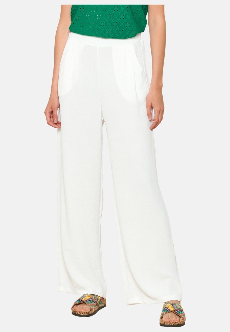 Weiße, fließende weit geschnittene Hosen aus leichtem Material. Verfügt über Vordertaschen und einen hohen Taillenbund. Die Schuhe sind bunt und mit Mustern versehen.