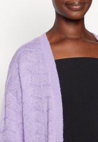 Lavendel gestrickte Strickjacke mit strukturiertem Muster, offenem Vorderteil und weiten Ärmeln, über einem schwarzen trägerlosen Top. Weicher, leichter Stoff.