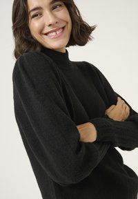 Pull en maille noir avec un col haut, des manches bouffantes et des bords côtelés. Texture douce, silhouette ajustée et design minimaliste.