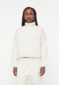 Svetlo béžová skrátená bunda z mäkkej fleece, s vysokým golierom, zipsom a detailom loga na hrudi; v kombinácii s ladícími nohavicami.