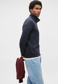 Felpa blu navy con zip e collo alto, tessuto testurizzato e polsini a coste, abbinata a jeans azzurri e un indumento bordeaux in mano.
