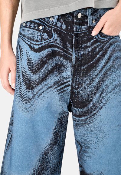 Blue denim pants featuring a swirling black print, with silver hardware and a relaxed fit. Textured appearance resembling brush strokes.