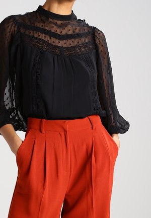 Femme portant un chemisier noir transparent à pois avec des détails en dentelle et un pantalon plissé taille haute couleur rouille avec les mains dans les poches.