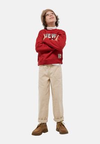 Sweatshirt rouge avec imprimé "NEW YORK", pantalon en velours côtelé crème et bottes marron à lacets. Le mannequin porte un bonnet en tricot beige et un col roulé blanc.