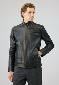 Man in een zwarte leren jas met rits en hand in de zak, kijkend naar de zijkant tegen een effen lichte achtergrond.