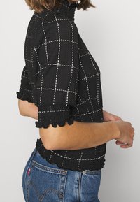 Sort tekstureret top med hvidt rutenetmønster, smockkrave og yndige ærmer, parret med blå jeans.